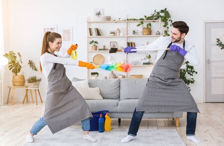 Cleaning with joy. Guy and girl in aprons have fun, fight with spray, brush and broom.の写真素材