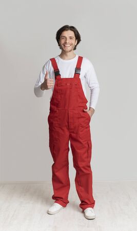 Smiling young laborer posing in work overalls and showing thumb up over light studio background, full-length shot, copy spaceの写真素材