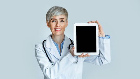 Smiling woman doctor showing tablet with blank screen, isolated on light background, studio shotの写真素材