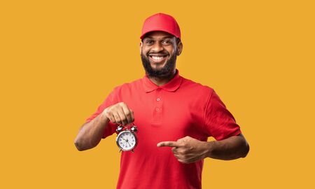 On-Time Delivery. Black Courier Pointing Finger At Clock Smiling To Camera Standing On Yellow Studio Background.の写真素材