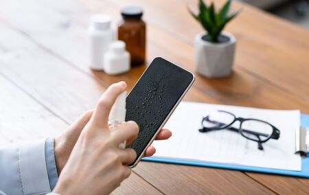 Fight off viruses and germs. Women hands disinfect phone with antiseptic. Pills, glasses and tablet on doctor deskの写真素材