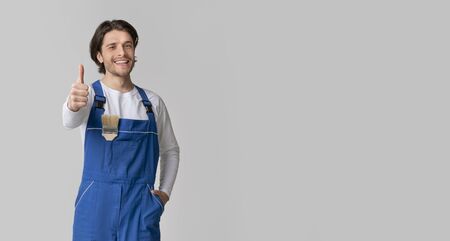 Repair And Renovation. Smiling handyman with paintbrush in pocket showing thumb up, posing over light background in studio, panorama with copy spaceの写真素材