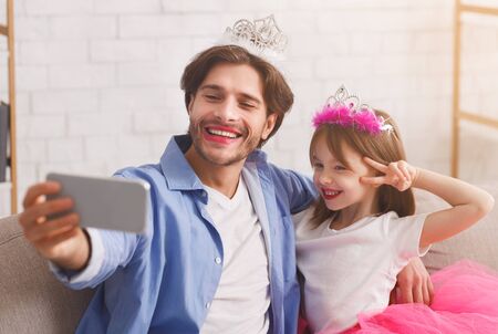 Happy father and daughter wearing as princesses taking selfie at home, happy moments togetherの写真素材