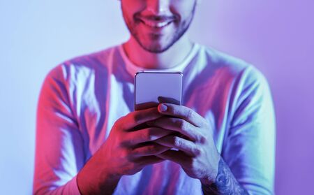 Millennial man is online. Smiling guy is typing in smartphone, cropped in neon, studio shotの写真素材