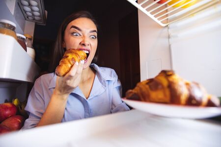 Ripped off diet. Hungry woman eating croissant from fridge, view from insideの写真素材