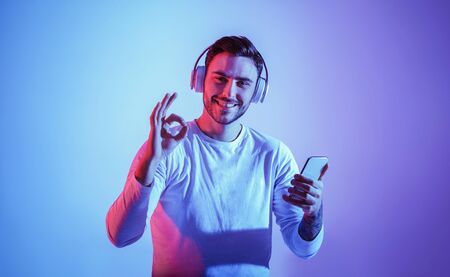 Symbol with hands is excellent. Smiling millennial guy with large wireless headphones expresses joy and holds smartphone, free spaceの写真素材