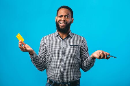 Worrying About Money. Confused black man holding credit card and smartphone, shrugging. Copyspace,...