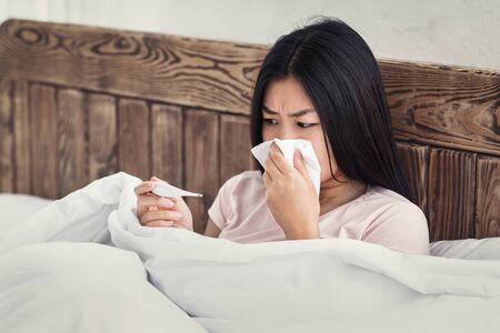 Influenza. Sick Chinese Woman Having Fever Holding Thermometer And Blowing Nose In Tissue Sitting In Bed Indoorsの写真素材