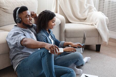 Relaxed Black Guy Listening Music In Wireless Headphones At Home While His Girlfriend Working On...