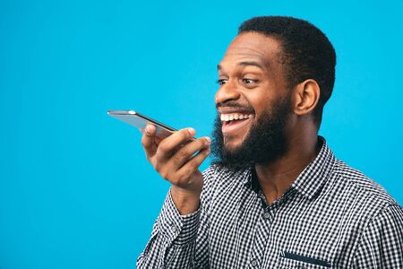 Happy young black man using voice assistant on smartphone, isolated over blue studio wall, copyspaceの写真素材