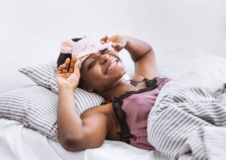 Smiling african american woman woke up, removes mask for sleeping, in bed on light backgroundの写真素材