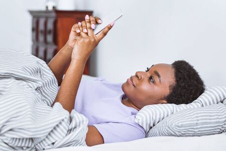 Chat in bed. African american woman typing on smartphone in bed in interior of bedroom, free space