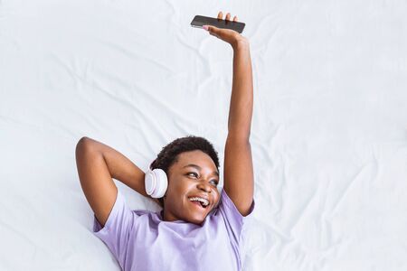 Enjoy favorite music. African american girl lies in bed with smartphone, listened song in headphones, free space, top viewの写真素材