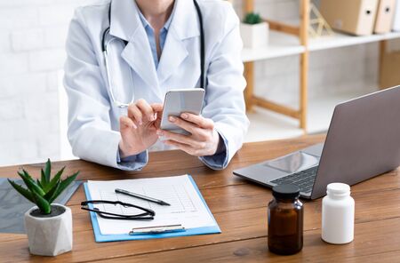 Remote online patient consultation. Female doctor typing on phone, on table with pills, tablets and...