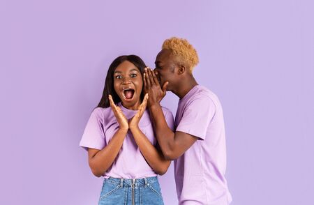 Handsome black man whispering secret or interesting gossip to womans ear on lilac background
