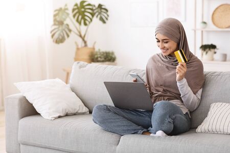 Online Shopping. Smiling muslim woman in hijab using smartphone, laptop and credit card at home, purchasing goods in internet, free spaceの写真素材