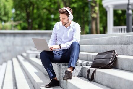 Handsome businessman with headphones and laptop communicating online while sitting on stairs in city...