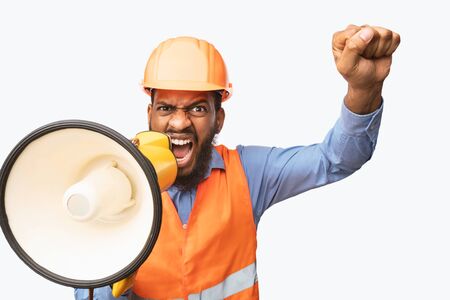 Emotional Black Construction Worker Shouting In Megaphone Looking At Camera And Shaking Fist Over White Background. Studio Shotの写真素材