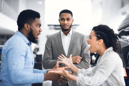 Too Expensive Car. Dealer Looking At African Spouses Having Quarrel About Buying Vehicle Standing In Dealership Store. Selective Focusの写真素材
