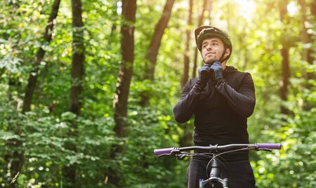 Concentrated guy cyclist locking helmet before ride in summer forest, staying with bike in sun...