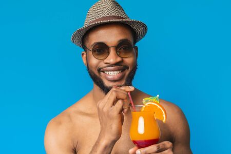 Handsome black guy in stylish hat enjoying fruit non-alcoholic cocktail on blue studio background,...