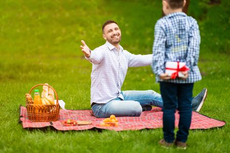 Fathers Day celebration. Little boy hiding gift for his dad behind his back on picnic in parkの写真素材