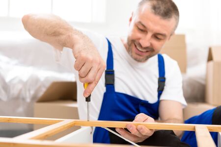 Carpentry And Furniture Assembly. Carpenter Making Wooden Shelf Using Screwdriver, Wearing Blue...