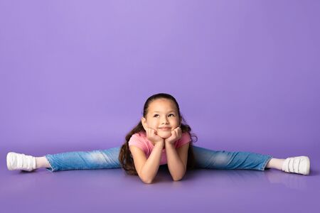 Stretching Concept. Cute little asian girl doing gymnastic splits on pastel purple studio background, copy spaceの写真素材