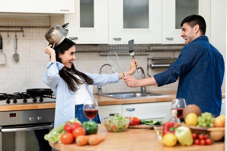 Protect Yourself. Playful man and woman fighting with kitchen supplies, having fun together at homeの写真素材