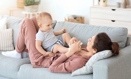 Young mother relaxing on couch with her adorable infant baby son, toddler boy resting on sofa with...