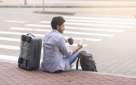 Male tourist sitting near airport terminal using smartphone and enjoying takeout coffee, free spaceの写真素材