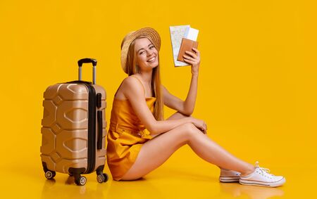 Travel Insurance Concept. Happy Young Girl Sitting On Floor With Suitcase And Tickets, Ready For Vacation Trip, Yellow Studio Backgroundの写真素材
