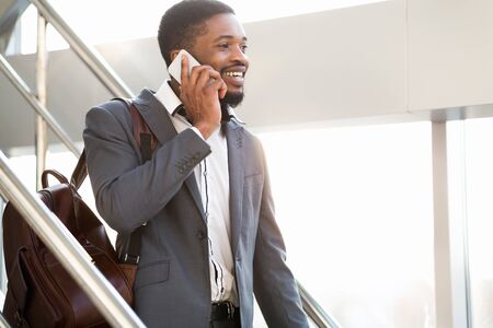 Smiling African Man Talking On Cellphone Walking With Backpack Down The Stairs In Airport Indoors....