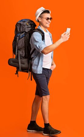 Smiling hiker using trakking application on mobile phone while walking over orange studio backgroundの写真素材