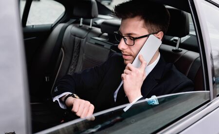 Hurry Up. Focused worker in eyewear making phone call, looking at luxury wristwatch, sitting in taxi