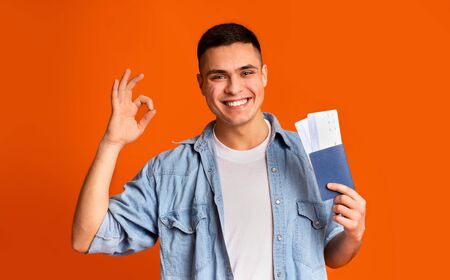 Happy young man with passport and flight tickets showing okay gesture over orange backgroundの写真素材