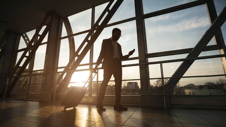 African   Entrepreneur On Business Travel Using Phone Walking With Baggage In Airport Terminal. Panorama, Free Spaceの写真素材