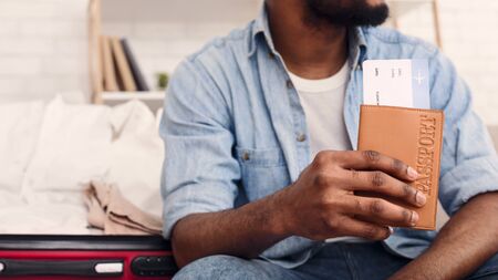 Flight Tickets. Unrecognizable African Man Holding Boarding Pass And Passport Sitting With Suitcase At Home.の写真素材
