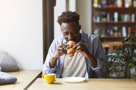 Funky African   guy having breakfast in cafe, eating delicious croissant with coffeeの写真素材