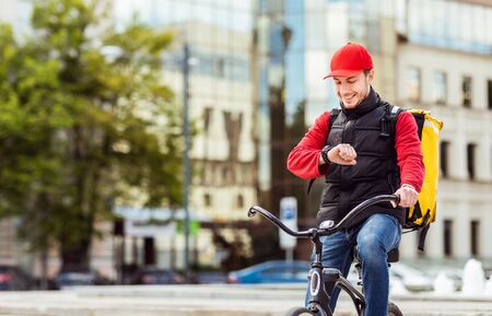 On-Time Food Delivery. Courier On Bike Checking Time On Wristwatch Delivering Meals From Cafes And Restaurants In City. Copy Spaceの写真素材