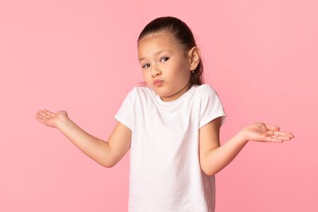 I Dont Know. Cute asian girl shrugging shoulders with spreaded hands, pastel pink studio wall, copy...