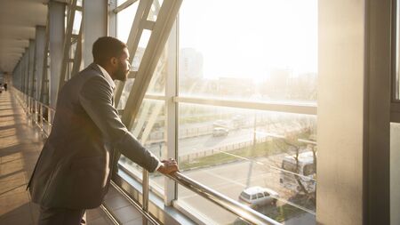 Black Businessman Standing In Airport Terminal Waiting For Flight Looking Through Window At City....