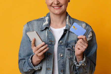 Easy Payments. Smiling middle-aged woman using smartphone and credit card for online money transfers, yellow background, cropped imageの写真素材