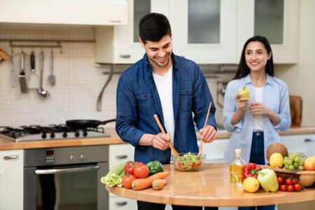 Healthy Eating Concept. Handsome man stirring fresh vegetable salad in glass bowl, girl standing in the blurred backgroundの写真素材