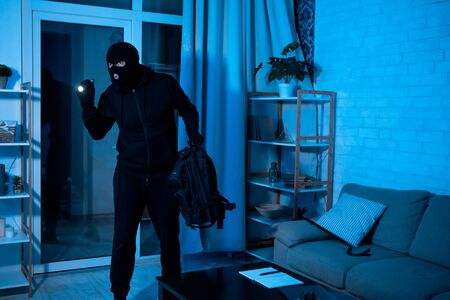 Robbery Concept. Disguised burglar standing in apartment or office, using flashlight, searching...
