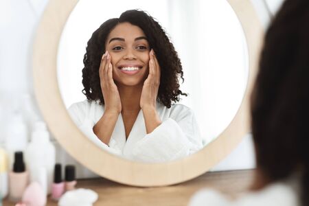 Young black girl caring of her beautiful face, enjoying touching soft skin, reflecting in mirror in...