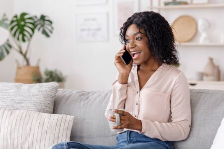 Spare time at home. Joyful black woman talking on cellphone and enjoying coffee while relaxing on...