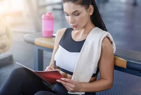 Beautiful millennial girl writing her workout plan in notebook at fitness clubの写真素材