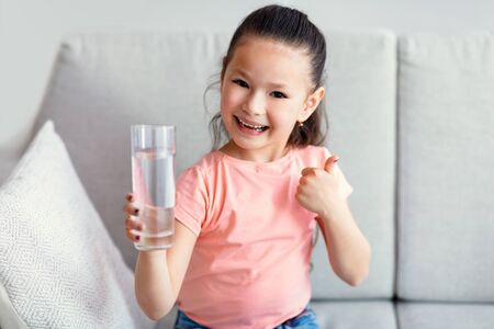 Healthy Hydration. Cute Little Asian Girl Holding Glass Of Water Gesturing Thumbs Up Sitting On Couch At Homeの写真素材