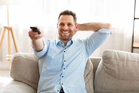 Relax Concept. Smiling calm man in shirt watching TV on the couch at home, holding remote control,...